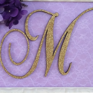 Op de afbeelding: Een ingelijst decoratief stuk met de letter "M" bedekt met goudglitter tegen een lavendelkleurige achtergrond met kantpatroon. De lijst is wit en paarse bloemen sieren de linkerbovenhoek. Geschikt voor interieurdecoratie.