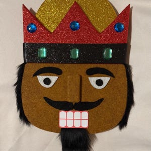 May include: A nutcracker face decoration made of brown felt with black faux fur hair and a beard. It features a red glitter crown with blue and green plastic gems, a gold glitter hat top with a red star, white felt eyes, and a white toothy mouth.
