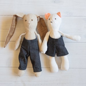 May include: Two handmade plush toys, a bunny and a cat, wearing denim overalls. The bunny is beige with brown ears and the cat is white with orange ears.