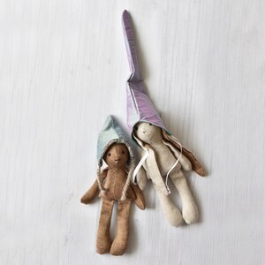 May include: Two handmade fabric dolls, one brown with a blue and green hat and the other beige with a purple hat. The dolls are lying on a white wooden surface.