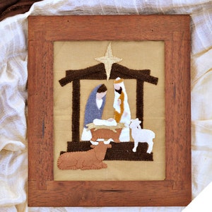 May include: A framed felt Christmas nativity scene with a brown wooden frame. The scene depicts Mary, Joseph, and baby Jesus in a manger with a star above them. There are also two animals, a cow and a sheep, in the scene.