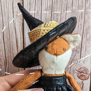 May include: A miniature witch hat tutorial featuring a small stuffed fox wearing a black witch hat with a gold trim. The hat is decorated with gold embellishments. The background is a wooden surface.