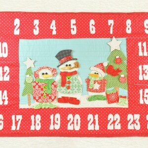 May include: A red felt advent calendar with white numbers 1 through 24. The calendar features a scene with three snowmen and a Christmas tree.
