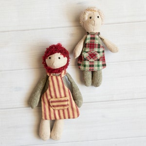 Könnte beinhalten: Zwei handgefertigte Stoffpuppen mit rot-weiß gestreiften und karierten Schürzen. Die Puppen haben beige Stoffkörper und Gesichter mit schwarzen Augen und roten Wollhaaren.