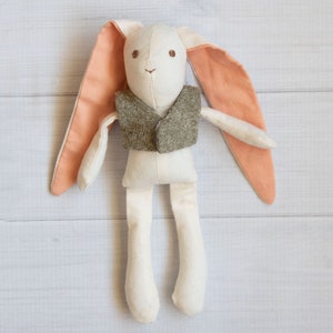 May include: A white fabric bunny toy with peach-colored ears and a brown tweed vest. The bunny has embroidered eyes and a nose.