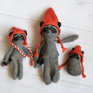 May include: Three grey fabric raccoon toys wearing orange knitted hats. The toys are standing on a white wooden surface.