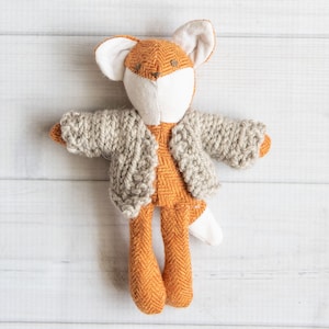 Könnte beinhalten: Ein Plüschfuchs-Spielzeug, das einen gestrickten Cardigan trägt. Der Fuchs ist orange und weiß mit einem langen Schwanz. Der Cardigan ist grau und hat ein Zopfmuster.