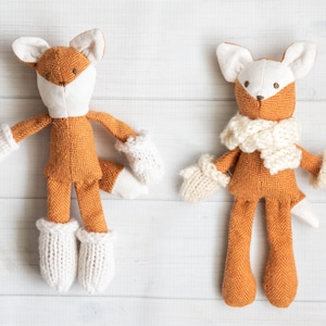 Könnte beinhalten: Zwei braune und weiße Plüsch-Füchse mit gestrickten weißen Fäustlingen und einem gestrickten weißen Schal. Der Fuchs links hat einen weißen Schwanz und der Fuchs rechts ein weißes Gesicht.