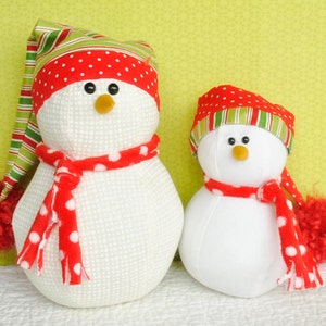 Peut inclure: Deux décorations de bonhomme de neige blanches avec des chapeaux rayés rouge et vert et des écharpes rouges à pois blancs. Le plus grand bonhomme de neige a un pompon rouge sur son chapeau.