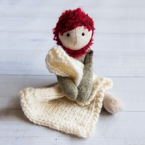 Könnte beinhalten: Eine handgemachte Puppe mit roten Locken, die ein grünes und braunes Outfit und eine weiße Strickdecke trägt.