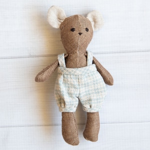 May include: A brown plush toy bear wearing blue and white checkered overalls. The bear has large ears and a stitched nose.