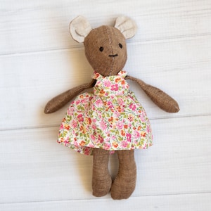 May include: A brown plush toy with large ears and a floral print dress. The toy has a simple face with two black eyes and a stitched mouth.