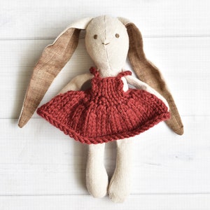 Könnte beinhalten: Eine handgemachte Plüsch-Hasenpuppe, die ein gestricktes burgunderfarbenes Kleid trägt. Die Puppe hat lange braune Ohren und ein weißes Gesicht mit gestickten Augen und einem Mund.