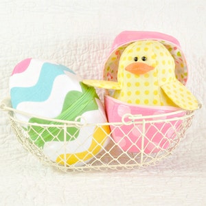 May include: An Easter display featuring a yellow, polka-dotted duckling toy in a pink eggshell container, placed in a white wire basket. The basket also contains colourful Easter eggs.