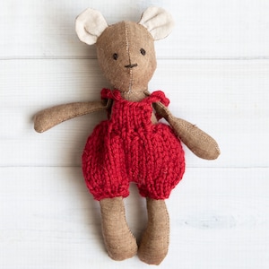 Könnte beinhalten: Ein brauner, gestrickter Plüschbär, der eine rote, gestrickte Latzhose trägt. Der Bär hat weiße Filzohren und schwarze gestickte Augen.