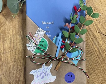 May include: A wrapped gift with a blue notebook that reads "Blessed is he who". The gift is wrapped in brown paper and decorated with stickers, a pen, and a sprig of greenery. A red, green, and white string is tied around the gift.