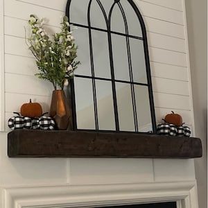 Puede incluir: Una repisa de chimenea de madera marr&oacute;n oscuro con dos peque&ntilde;as calabazas a cuadros naranja y blanco a cada lado. Un jarr&oacute;n de cobre con flores blancas se encuentra en la repisa frente a un espejo de ventana arqueado negro.