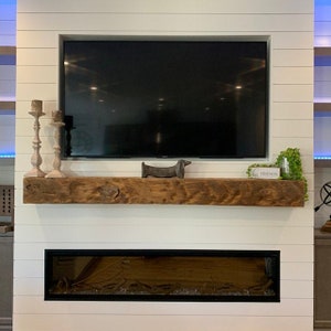 Puede incluir: Una pared blanca con un televisor de pantalla plana montado sobre una repisa de madera. La repisa tiene una peque&ntilde;a figura de dachshund de madera y un letrero que dice "FRIENDS". Debajo de la repisa hay una chimenea el&eacute;ctrica enmarcada en negro.