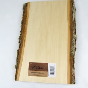 May include: A natural wood plank with live edge, featuring a light brown color and a rough, textured surface. The plank has a label that reads "Wilson BASSWOODplank (medium)" and a website address.