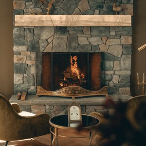 Puede incluir: Una chimenea de piedra con un fuego crepitante en su interior. La chimenea tiene una rejilla de lat&oacute;n y una repisa de madera encima. Dos sillas est&aacute;n colocadas frente a la chimenea, con una mesa peque&ntilde;a entre ellas. La mesa tiene una tapa de vidrio negro y un marco de metal dorado. Un peque&ntilde;o cuadro enmarcado se encuentra sobre la mesa.