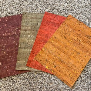 May include: Four rectangular cork sheets in shades of burgundy, olive green, red-orange, and golden-orange. The cork has a textured, speckled appearance, with variations in color and pattern. The sheets are arranged on a neutral-colored carpet.
