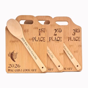 May include: Three rectangular bamboo cutting boards with handles, each engraved with 1st, 2nd, and 3rd Place and 2026 BAC Chili Cook-Off. Three wooden spoons are placed diagonally across the boards, each engraved with 2026 BAC Chili Cook-Off.