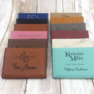 May include: A collection of leather business card holders in various colors, including brown, pink, and teal. Each holder is engraved with text and logos, such as "Your 901 Connection" and "Kreischer Miller". The holders are rectangular with stitched edges.