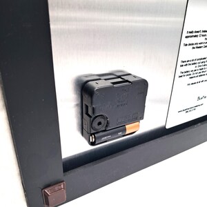 May include: The back of a wall clock featuring a black plastic battery-operated quartz movement mechanism with an AA battery inserted. A silver metal plate is attached to the black frame, displaying printed text instructions and the BlueFire logo.