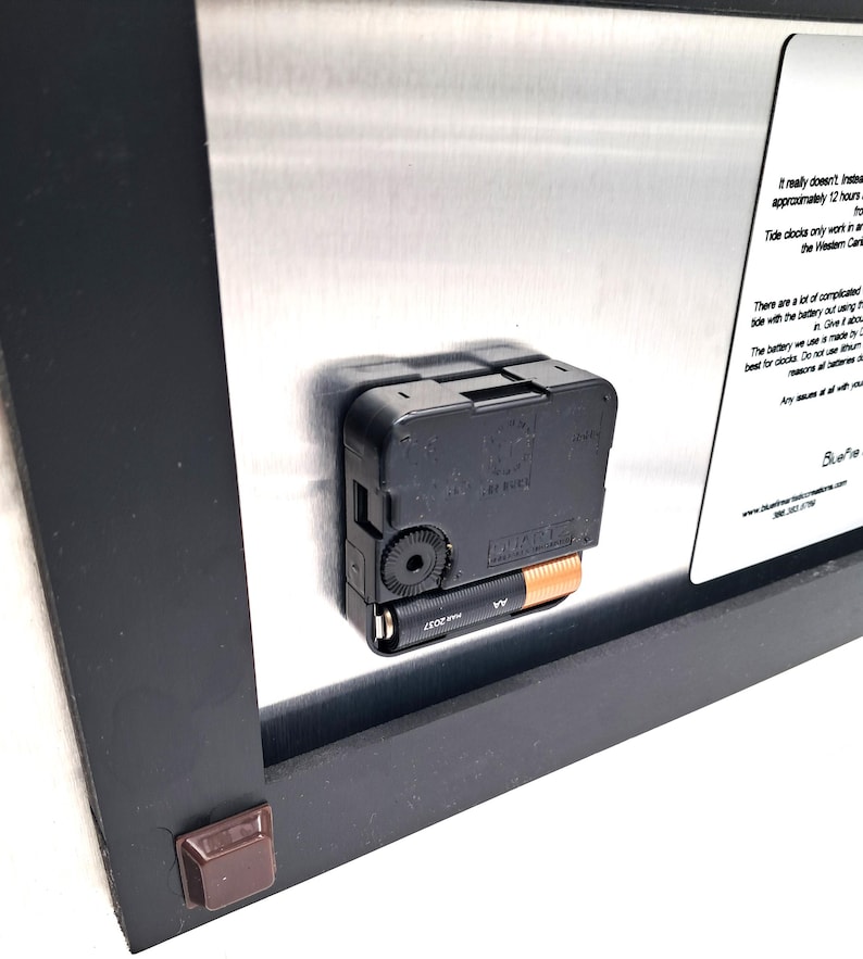 May include: The back of a wall clock featuring a black plastic battery-operated quartz movement mechanism with an AA battery inserted. A silver metal plate is attached to the black frame, displaying printed text instructions and the BlueFire logo.