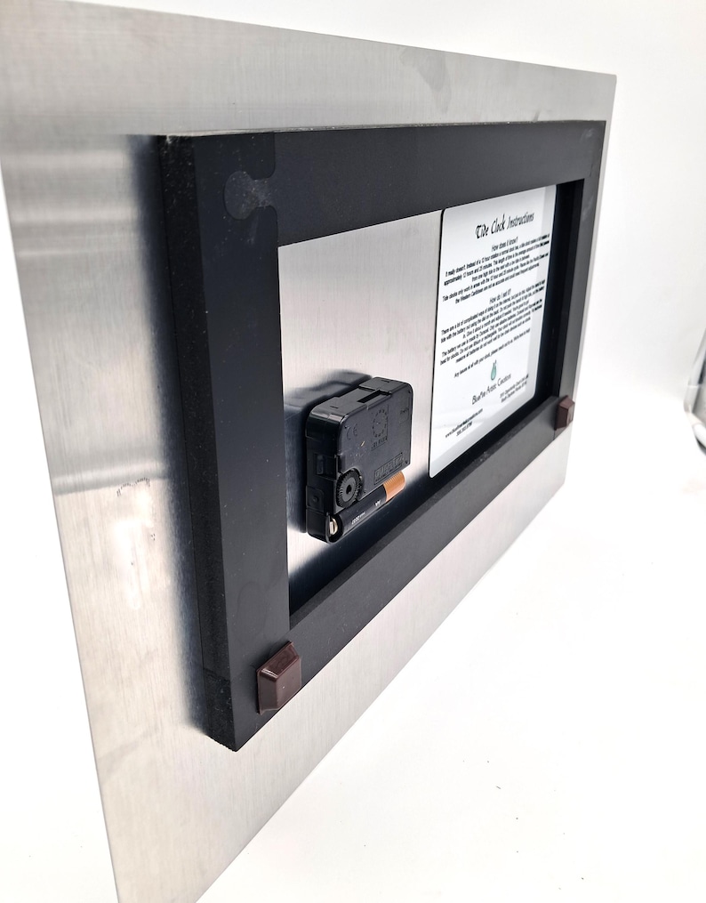 May include: The back of a rectangular metal clock featuring a black plastic frame, a central battery-operated clock mechanism with a single AA battery inserted, and a white instructional label titled The Tide Clock Instructions.