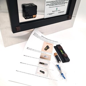 May include: The back of a rectangular metal clock with a black battery-operated mechanism and a white information label. Included are wall hanging instructions, a black level tool, a single AA battery, and a plastic bag with screws and anchors.