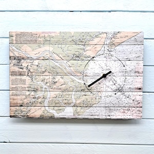 May include: A rectangular wooden wall clock featuring a nautical map design. The clock face displays a map of the Savannah River area, with black clock hands and text indicating tide levels. The clock is mounted on a light blue wooden surface.