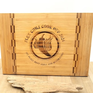 May include: A brown bamboo cutting board with a laser-engraved design. The design features a chili pepper with flames and the text "Flex Chili Cook Off 2024 1st Place Best Chili and Decoration".