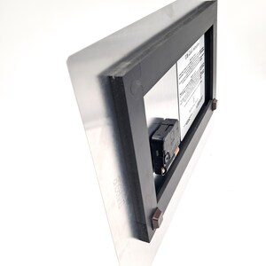 May include: The back of a rectangular metal wall clock featuring a black plastic frame, a black battery-operated clock mechanism, and a white instructional label with black text. Four small brown square spacers are attached to the frame corners.