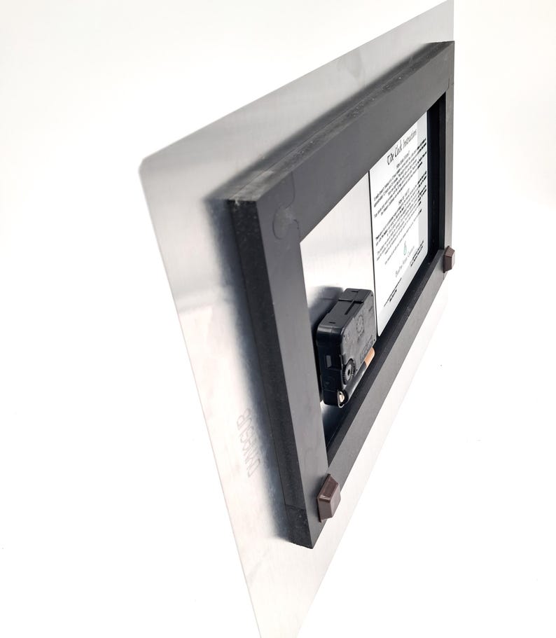 May include: The back of a rectangular metal wall clock featuring a black plastic frame, a black battery-operated clock mechanism, and a white instructional label with black text. Four small brown square spacers are attached to the frame corners.