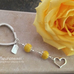 May include: A silver heart-shaped keyring with a small tag that says "Fleurenasci bringing flower essence to life". The keyring has two yellow beads attached to it.