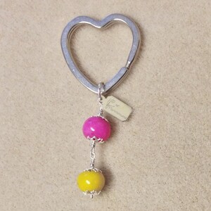 May include: A silver heart-shaped keyring with a pink and yellow bead charm. The charm has a silver tag with the text "Love is Handmade".