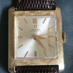 May include: A gold-toned Omega Automatic wristwatch with a square face and a brown leather band.