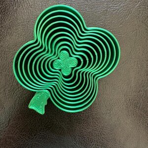 May include: A green plastic clover-shaped cookie cutter set. The set includes multiple sizes of the clover shape, nested within each other. The cutter has a leaf detail on the side. The set is designed for baking and crafting.