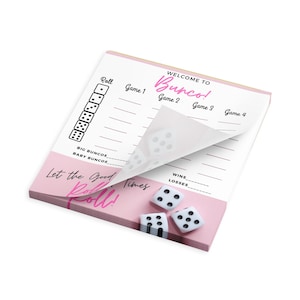 May include: A pink and white Bunco score pad with the words "Welcome to Bunco!" and "Let the Good Times Roll!" printed on it. Three white dice with black dots are next to the pad. The pad is for keeping score during a Bunco game.
