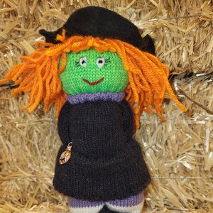 May include: A knitted witch doll with orange hair, a black hat, and a black dress. The doll is wearing purple and white striped socks and black shoes. The doll is sitting on a bed of straw.