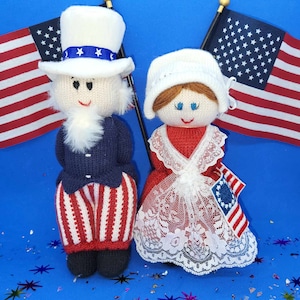 May include: Two knitted dolls dressed in red, white, and blue patriotic clothing. The doll on the left is wearing a blue jacket and red and white striped pants. The doll on the right is wearing a white dress with red and white lace trim. Both dolls are holding American flags.
