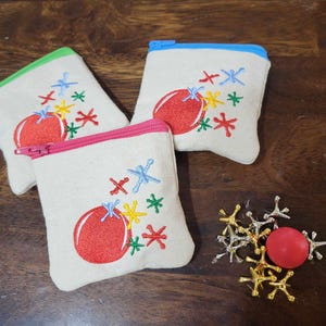 May include: Three small fabric pouches with red, blue, and green zipper pulls. Each pouch has a white background with an embroidered red circle and colorful stars. The pouches are on a wooden surface with a pile of silver and gold jacks and a red jack.
