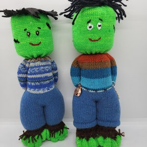 May include: Two green knitted plush toys with black hair, blue pants, and brown shoes. The toys have smiling faces with black eyes and red mouths.