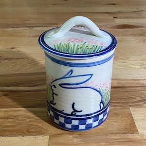 以下が含まれることがあります： 手描きのデザインが施された、蓋付きの陶器のジャー。ジャーは、青いウサギのイラストとピンクの花柄のアクセントが付いた白いベースです。蓋には白いハンドルと青い縁が付いています。ベースは青と白のチェック柄です。