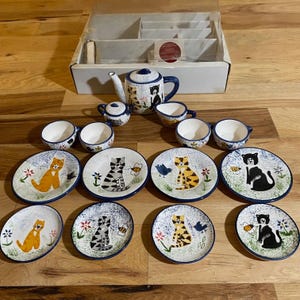 May include: A vintage ceramic tea set featuring cat designs. The set includes a teapot, sugar bowl, creamer, cups, and saucers. Each piece is decorated with cat illustrations, flowers, and a blue rim. The set is displayed in its original box.