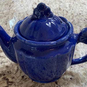 May include: A cobalt blue ceramic teapot with a matching lid. The teapot has a rounded body, a curved spout, and a handle with a floral design. The lid is topped with a decorative floral knob.