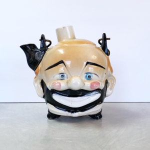 May include: A ceramic teapot shaped like a clown's head with a black and white painted face, blue eyes, and a wide smile. The teapot has a black and white striped collar and black shoes.