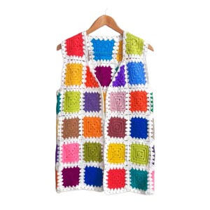 May include: A colorful, handmade crochet vest with a patchwork design. The vest features squares in various colors, including red, blue, green, yellow, and purple, all outlined in white. It hangs on a wooden hanger against a white background.