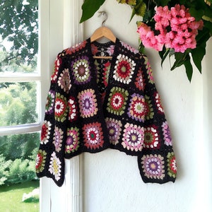 Puede incluir: Una chaqueta de punto de ganchillo negra con un patrón geométrico colorido. La chaqueta tiene una forma cuadrada y está hecha con una variedad de colores, incluyendo rosa, rojo, verde y morado.